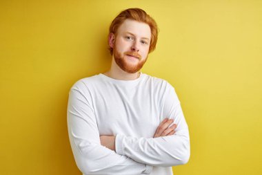 Kendine güveni tam, kızıl saçlı, kollarını kavuşturmuş beyaz gömlekli, sarı arka planda yalnız bir adam portresi.