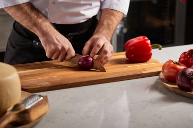 Aşçı erkek, restoranın mutfağında ahşap kesim tahtasıyla kırmızı soğan dilimliyor.