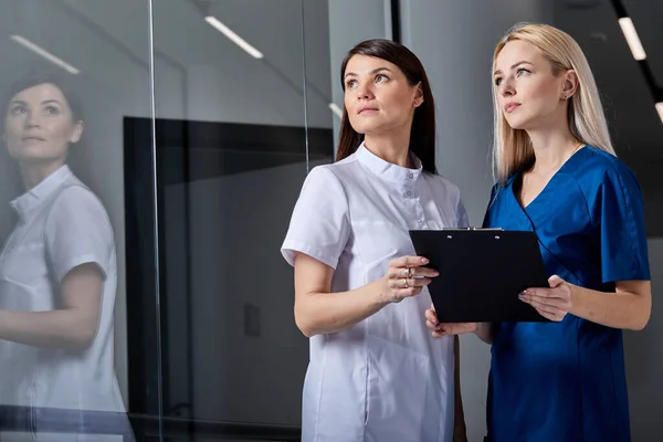 젊고 아름다운 두 명의 여성 의사들 이 옆을 잡고 있는 리스트 클립보드를 보고 있습니다. — 스톡 사진