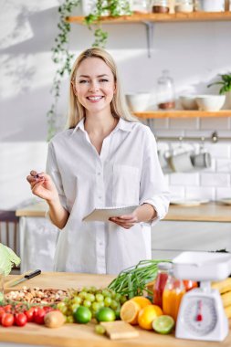 Kadın tarifleri panoya yazıyor. Masada taze meyve ve sebze var.
