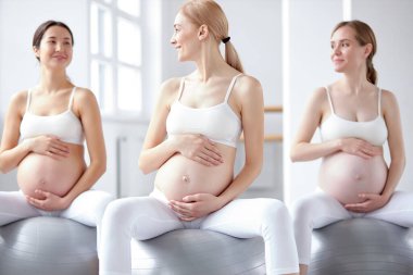 Güzel görünümlü, karnı çıplak hamile kadınlar perinatal jimnastik yaparken spor toplarının üzerine otururlar.