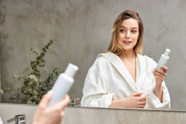 Bornozlu güzel bir manken krem maskeli beyaz bir şişe kullanıyor, banyoda aynanın önünde duruyor.