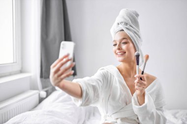 Havlu ve bornozlu neşeli kadın makyaj yaparken akıllı telefondan selfie çekiyor.