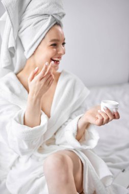 Beyaz bornozlu genç kadın yüzüne krem sür.