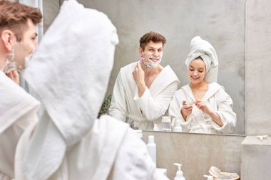 Bornoz ve havlu giymiş çekici bir kadın nemlendirici krem kullanırken erkek arkadaşı sakal tıraşı oluyor.