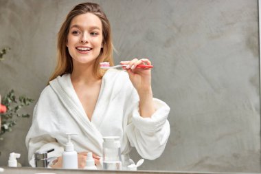 Bornozlu mutlu genç kadın temiz dişlerini fırçalayacak, banyodaki aynaya bakacak.