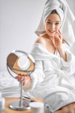 Beyaz havlu içinde güzel bir kadın yatak odasında dinleniyor ve küçük aynaya bakıyor. Güzellik prosedürleri uyguluyor.