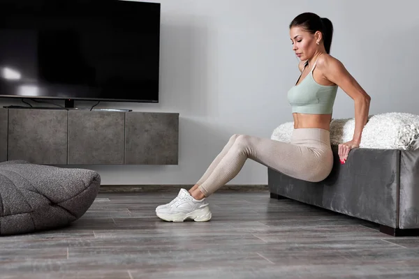 side view on fit female doing exercises near sofa, lifts body off the floor