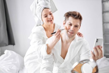 Bornoz ve havlu giymiş neşeli beyaz bir çift, erkek arkadaş yüzünde makyaj yapan bir kadın.