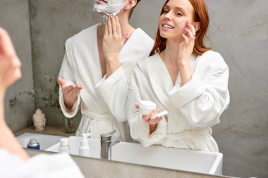 Beyaz tenli hoş bir çift yüz derisine bakar, kadın krem sürerken erkek sakal tıraşı olur.