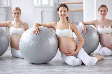 Genç hamile kadın spor yapıyor. Spor topu kullanıyor ve karnını tutuyor. Dinleniyor.