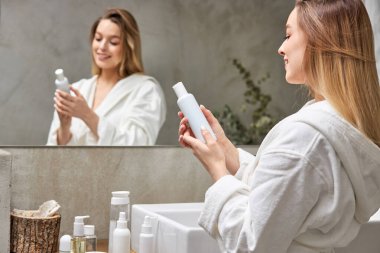 Bornozlu güzel bir manken krem maskeli beyaz bir şişe kullanıyor, banyoda aynanın önünde duruyor.