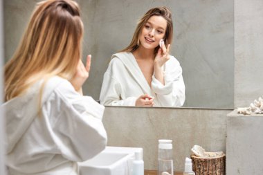 Bornozlu rahat kadın banyoda yüz ve boyun için nemlendirici serum uyguluyor.