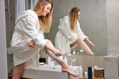 Banyoda bacaklarını traş eden, bornoz giyen genç bir kadın.