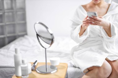 Sağlıklı ve rahat bir kadın akıllı telefon kullanarak oturur, banyodan sonra yatağında dinlenir.