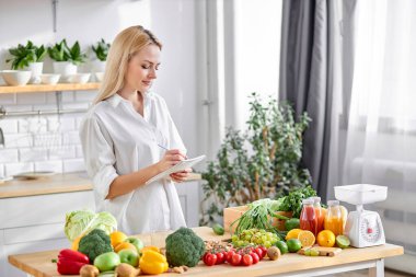 Beslenme uzmanı sarışın kadın, kendisi için diyet planı yazıyor. Meyve ve sebze dolu bir masa.