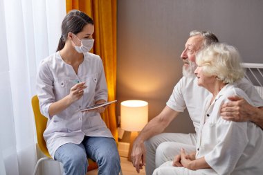 Yaşlı kadın doktor ev ziyaretinde, semptomları ve şikayetleri dinliyor, dijital tablete yazıyor.