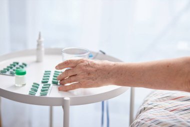 Yaşlı eller masada hap ve uyuşturucu alıyor, bir bardak su. Renkli tabletler taşıyan yaşlı bir kadının buruşuk elleri.