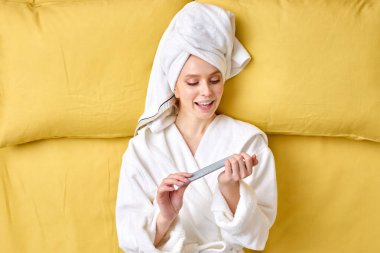 Bornoz ve havlu giymiş güzel bir kadın sabah ya da hafta sonları yatakta tırnaklarını kesiyor.