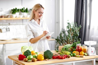 Kadın tarifleri panoya yazıyor. Masada taze meyve ve sebze var.
