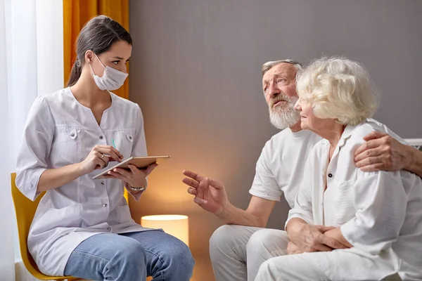 Jeune médecin en visite à domicile chez les personnes âgées, écoute les symptômes et les plaintes, écrit sur tablette numérique — Photo