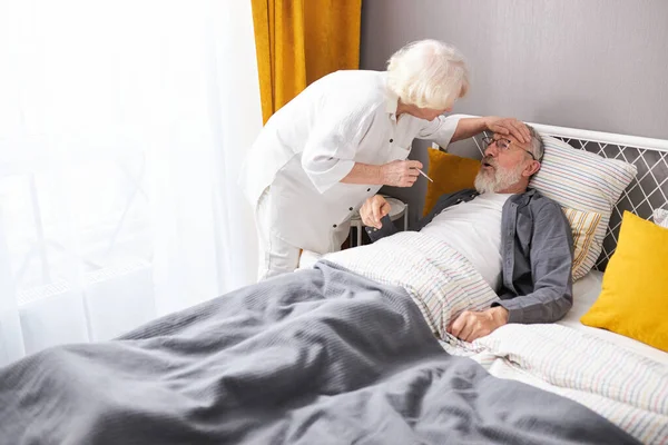 Femme prendre soin du mari malade, vérifier la température de la fièvre de l'homme âgé couché sur le lit à la maison — Photo
