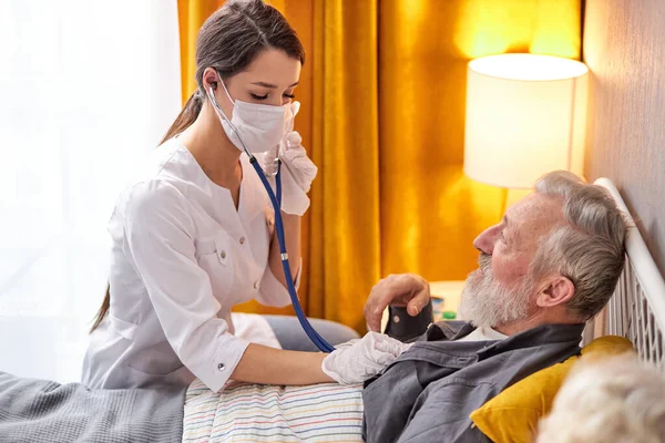 Jeune infirmière prendre soin d'un homme âgé sur un lit, infirmière écouter des sons dans le cœur ou les poumons en utilisant stéthoscope — Photo