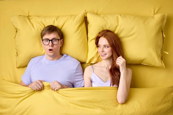 Caucasian couple lying in bed under duvet after first sex experience, man is surprised by skills of female in bed — Stock Photo, Image