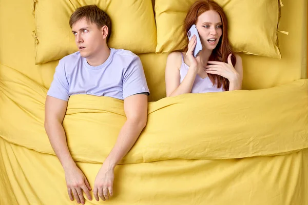 Junges Ehepaar im Bett, Frau telefoniert, Mann langweilt sich durch Gespräche — Stockfoto