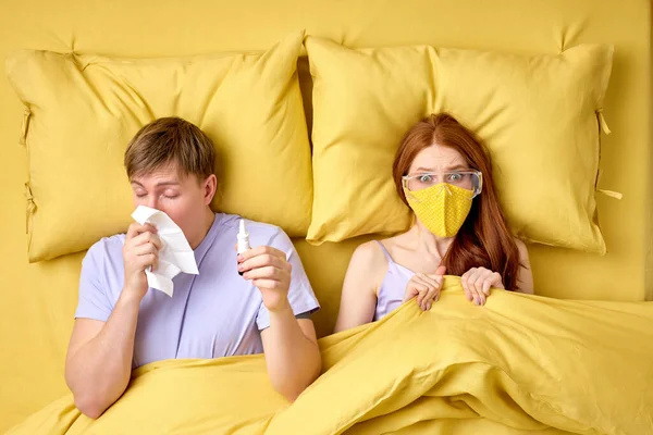 woman is afraid of catching coronavirus or flu infection, lying on bed with sick husband wearing mask and glasses