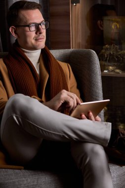 Gözlüklü adam rahat koltukta oturup bilgisayar tableti kullanıyor.
