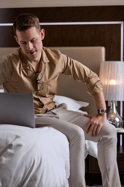 Giovane bel ragazzo sedersi sul letto in camera d'albergo, utilizzando il computer portatile — Foto Stock