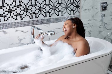 Siyah kadın banyo içinde köpüklerle banyo yapıyor, duş kabzasında şarkı söylüyor, eğleniyor.