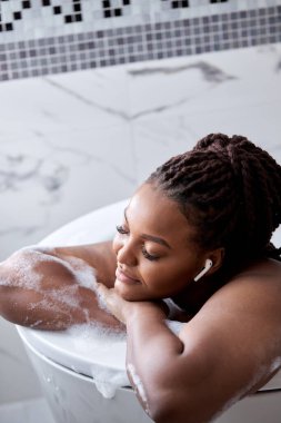 Rahat bir Afrikalı kadın köpükle banyo yapıyor, spanın, hijyenin tadını çıkarıyor ve müzik dinliyor.
