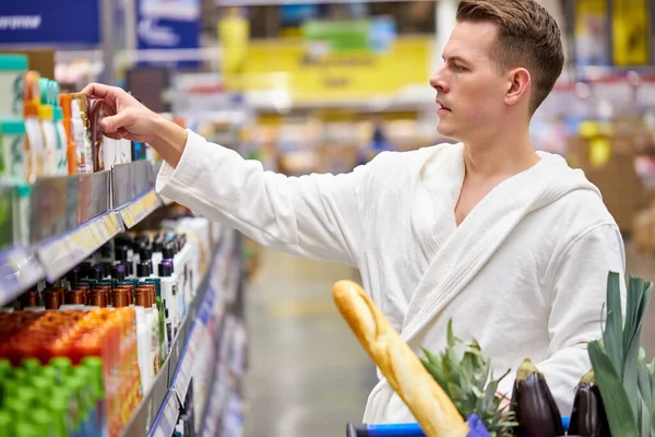 Καυκάσιος άνδρας με μπουρνούζι που ψάχνει για αφρόλουτρο της επιλογής του καλύτερου σαμπουάν στο σούπερ μάρκετ — Φωτογραφία Αρχείου