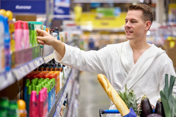 Side view on young male in bathrobe looking for shower gel of choosing the best shampoo in supermarket