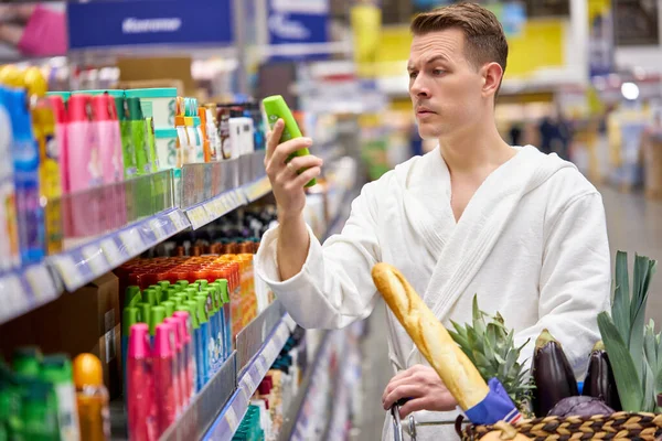 Скептик в халате читает информацию о шампуне, выбирая лучший в магазине — стоковое фото