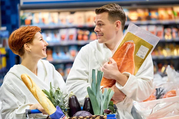 Молодая веселая пара в халатах веселится в супермаркете, выбирая сырую рыбу — стоковое фото