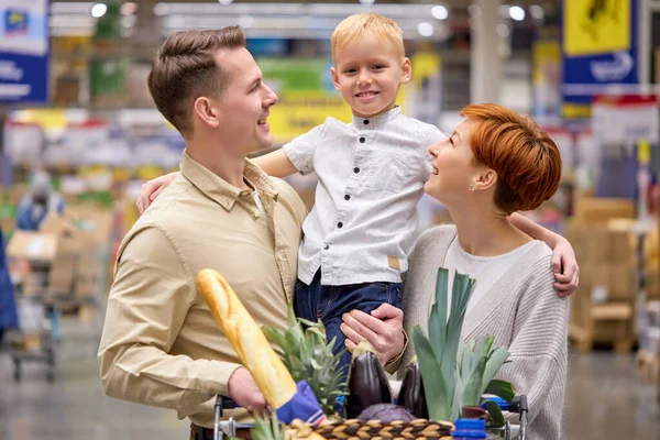 Retrato de familia joven y feliz comprando comestibles en el supermercado junto con un niño pequeño, sonriendo — Foto de Stock