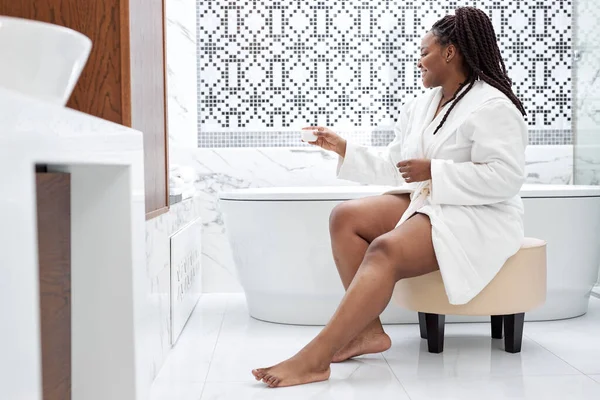 Афро-американка в халате проводит утро в ванной комнате, нанося увлажняющий крем на бритые и мягкие ноги — стоковое фото