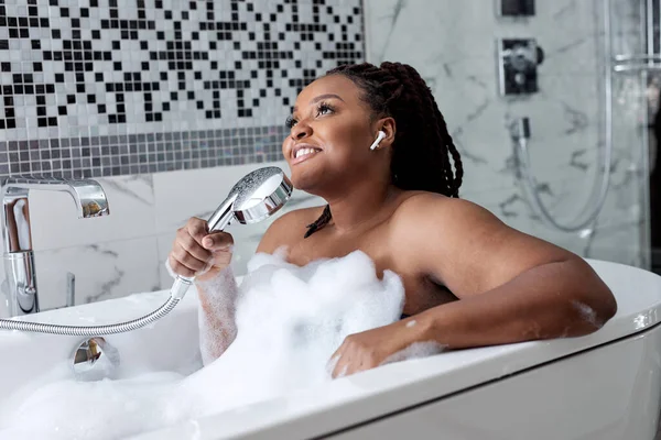 Красива усміхнена африканська жінка позує з мокрим волоссям у ванній кімнаті, розслабляючись у білій ванній — стокове фото