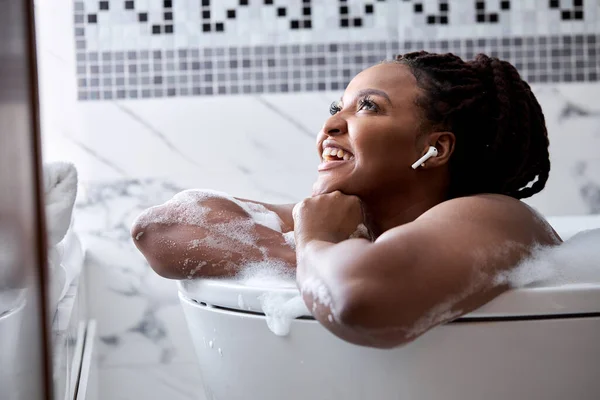 Мечтательная афро-американка лежит в ванной с пеной, отдыхает и слушает музыку в наушниках — стоковое фото