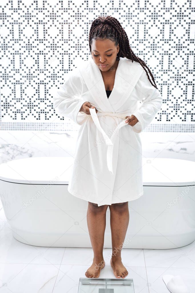 Young African American Female In Bathrobe Standing On Scales, Checking Weight