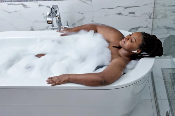 Afrikanerin im Bad. Badewanne mit Blasen und Schaum. Hautpflege und Behandlung im Whirlpool im Wellnessbereich — Stockfoto