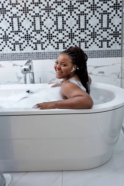 Hermosa mujer africana sonriente posando con el pelo mojado en el baño, relajándose en baño blanco en el baño — Foto de Stock