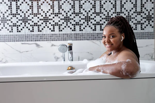 Linda mulher africana sorridente posando com cabelo molhado no banheiro, relaxando em banho branco no banheiro — Fotografia de Stock