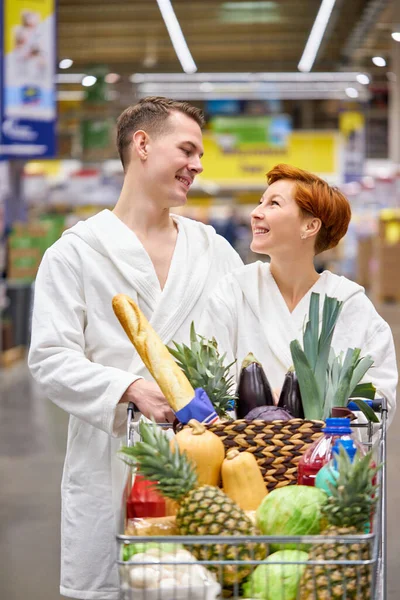 Jovem casal em roupões de banho no supermercado fazendo compras diárias andando com carrinho — Fotografia de Stock