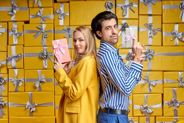 Adorable couple stand back to back posing at camera with gift boxes in hands