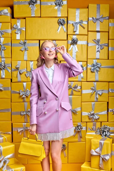 Señora atractiva en vestido rosa de moda y gafas de sol elegantes posando en la cámara, al lado de cajas de regalo — Foto de Stock