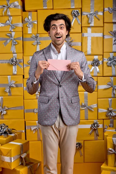 Esto es para ti. Hombre positivo en traje gris sosteniendo un certificado de regalo en las manos mientras está de pie lleno de cajas gtift — Foto de Stock